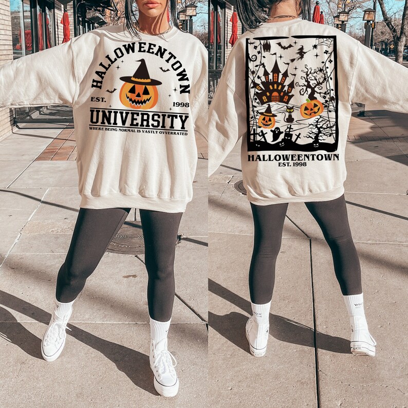 May include: Cream-coloured sweatshirt with "Halloweentown University" text and pumpkin graphic. The front design includes a pumpkin wearing a witch hat. The back features a Halloween-themed graphic with a haunted house, pumpkins, and bats. The text "EST. 1998" is included.