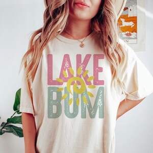 Op de afbeelding: Een vrouw draagt een cr&egrave;mekleurig T-shirt met de tekst "LAKE BUM" in roze, geel en blauw met een zon-graphic.