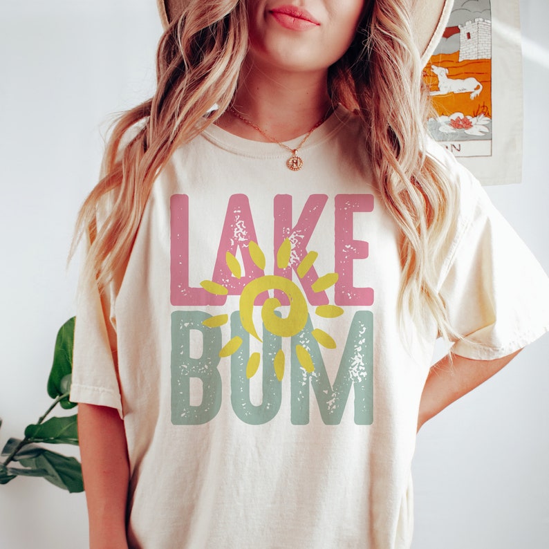 Op de afbeelding: Een vrouw draagt een cr&egrave;mekleurig T-shirt met de tekst "LAKE BUM" in roze, geel en blauw met een zon-graphic.