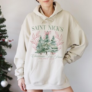 May include: Cream-colored hoodie with a front graphic featuring the text "SAINT NICK'S CHRISTMAS TREE FARM" above a watercolor illustration of Christmas trees. The design includes pink accents and a bow.