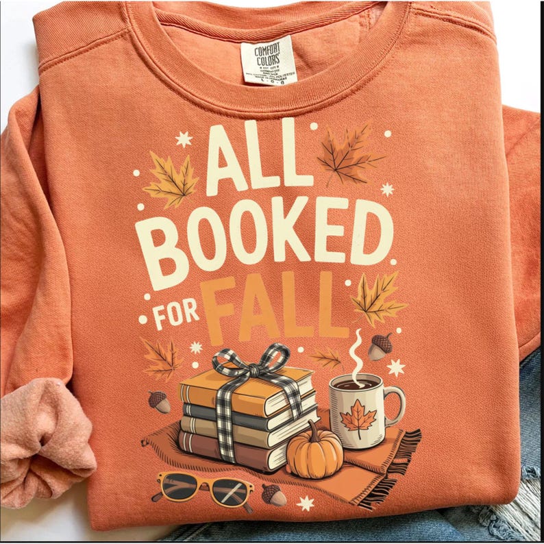 May include: Orange sweatshirt with the text "ALL BOOKED FOR FALL" and fall-themed graphics. The design includes books, a pumpkin, a coffee mug, and sunglasses. The sweatshirt is a Comfort Colors brand.