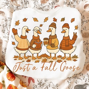 May include: White sweatshirt with a fall-themed graphic. The design features four cartoon geese wearing sweaters, hats, and scarves, surrounded by falling leaves. The text "Just a Fall Goose" is printed in a brown script font.
