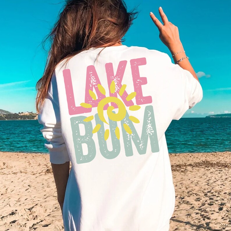 Op de afbeelding: Witte longsleeve met een roze en blauwe afbeelding met de tekst "LAKE BUM" met een gele zon in het midden.