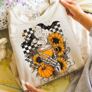 K&ouml;nnte beinhalten: Beiges Sweatshirt mit einem Halloween-Motiv. Das Design zeigt eine Kaffeetasse, Sonnenblumen, einen K&uuml;rbis, Flederm&auml;use und eine Skeletthand. Die Grafik ist vor einem karierten Hintergrund mit Sternen angeordnet.