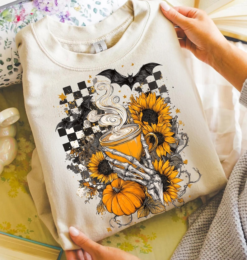 K&ouml;nnte beinhalten: Beiges Sweatshirt mit einem Halloween-Motiv. Das Design zeigt eine Kaffeetasse, Sonnenblumen, einen K&uuml;rbis, Flederm&auml;use und eine Skeletthand. Die Grafik ist vor einem karierten Hintergrund mit Sternen angeordnet.