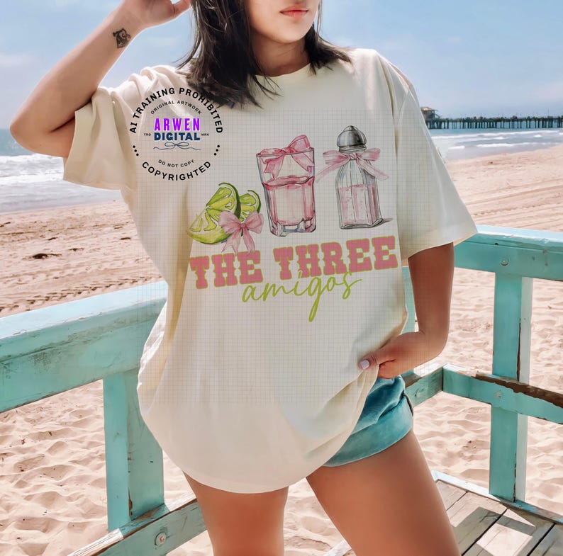 May include: Cream-colored t-shirt featuring a graphic of a cocktail, lime slices, and the text "THE THREE amigos" in pink and green. The design includes a cocktail glass with a pink bow and a bottle with a pink bow.
