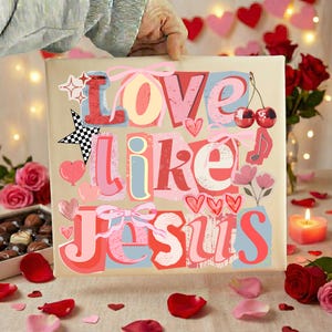 May include: A square canvas print with the words "Love Like Jesus" in a colourful, layered design. The artwork features hearts, bows, stars, and musical notes. The background is a neutral beige, with a hand holding the print.