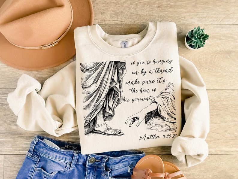 May include: A cream-colored sweatshirt with a black and white illustration of a hand reaching out from a robe. The text "if you're hanging on by a thread make sure it's the hem of his garment" is printed on the sweatshirt, along with the scripture reference "Matthew 9:20-22".