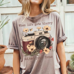 May include: A gray t-shirt with a vintage camera graphic and the text "When life gets blurry adjust your focus". The camera is surrounded by flowers.