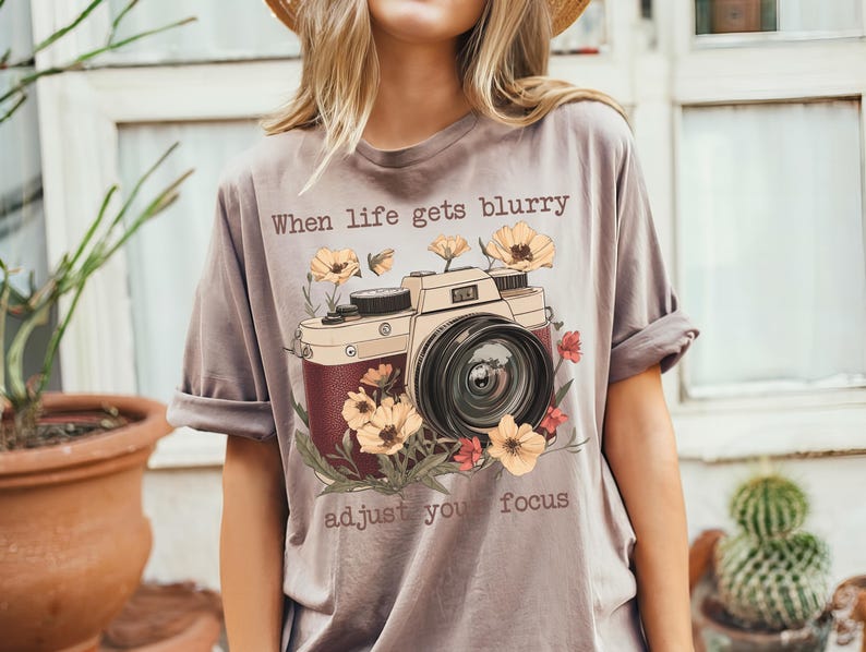 May include: A gray t-shirt with a vintage camera graphic and the text "When life gets blurry adjust your focus". The camera is surrounded by flowers.