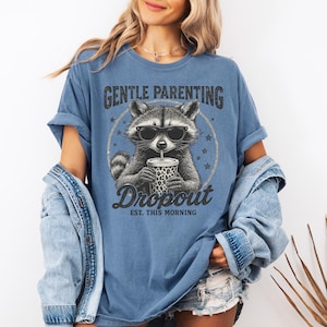 Puede incluir: Camiseta azul con un dise&ntilde;o de mapache con gafas de sol y una bebida. La camiseta tiene el texto "Gentle Parenting Dropout Est. This Morning" en una fuente desgastada. La camiseta es de corte holgado.
