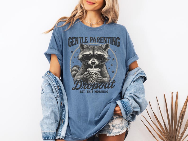 Puede incluir: Camiseta azul con un dise&ntilde;o de mapache con gafas de sol y una bebida. La camiseta tiene el texto "Gentle Parenting Dropout Est. This Morning" en una fuente desgastada. La camiseta es de corte holgado.