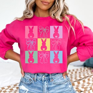 May include: A pink sweatshirt with a colorful pattern of bunnies and bows. The pattern is made up of nine squares, each with a different color and design.