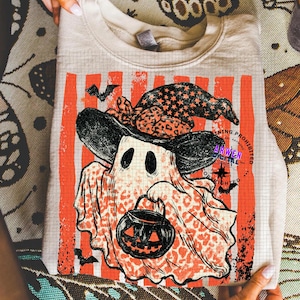 May include: A beige crewneck sweatshirt featuring a graphic of a ghost wearing a leopard print witch hat and holding a jack-o'-lantern bucket. The design includes vertical orange stripes, small bats, and the text Arwen Digital Copyrighting Prohibited.