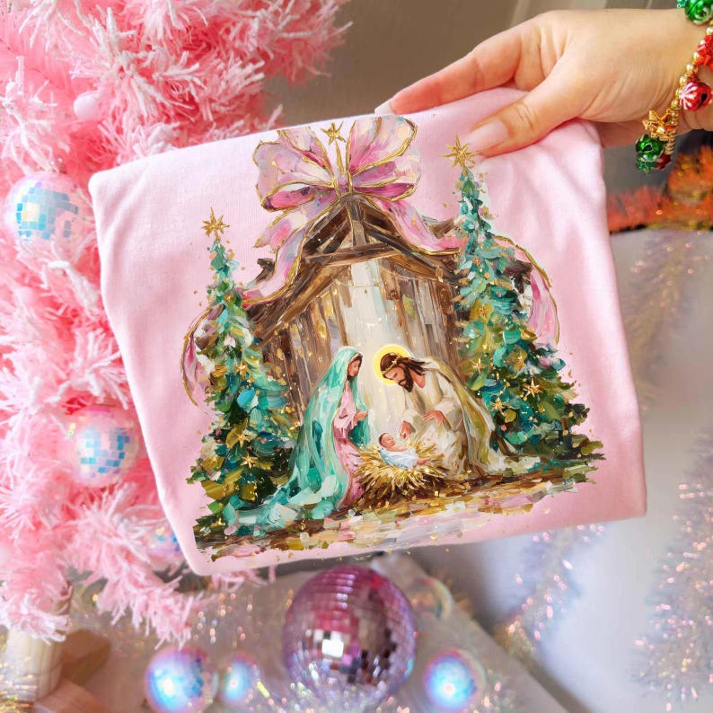 May include: A pink, square-shaped pillow featuring a Nativity scene with a baby Jesus, Mary, and Joseph. The image is framed by two Christmas trees and a large pink bow. The pillow is held by a hand.