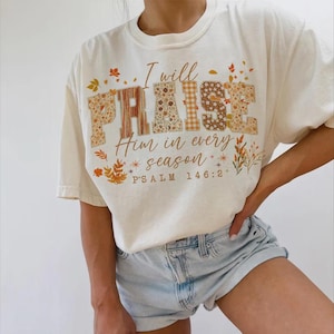May include: Cream-coloured t-shirt featuring the text "I will PRAISE Him in every season" and "PSALM 146:2" in brown. The word "PRAISE" is decorated with floral and plaid patterns. Autumn leaves and floral accents surround the text.