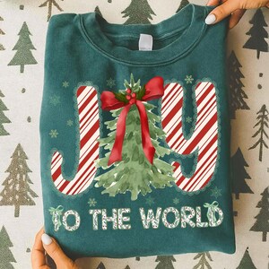 Puede incluir: Sudadera de color verde azulado con las palabras "JOY TO THE WORLD" en un dise&ntilde;o festivo. Las letras "J" e "Y" tienen rayas de bast&oacute;n de caramelo, y la "O" es un &aacute;rbol de Navidad de acuarela con un lazo rojo. Copos de nieve y una corona acent&uacute;an el dise&ntilde;o.