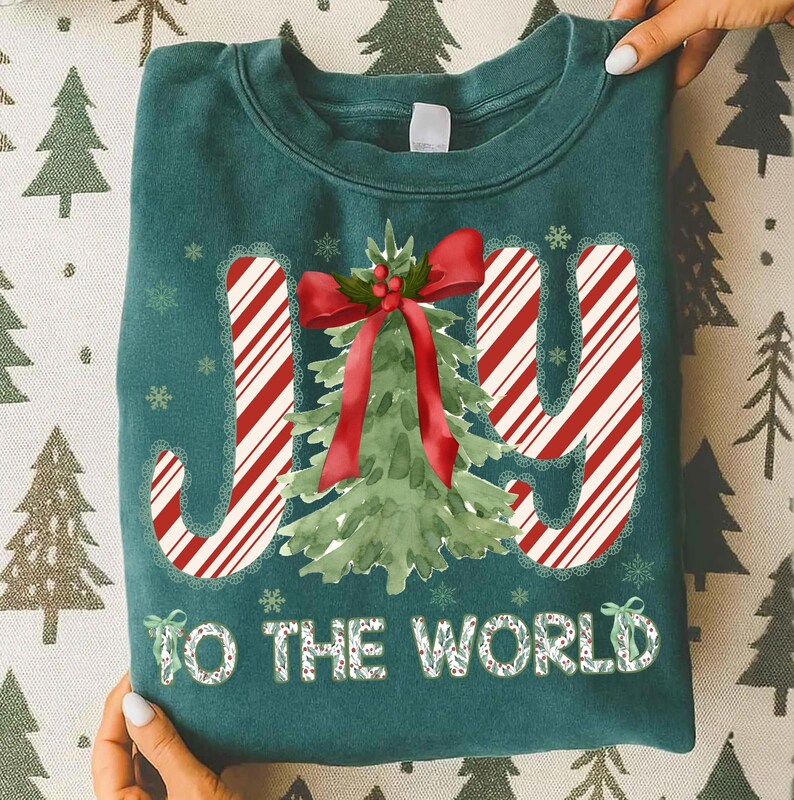 Puede incluir: Sudadera de color verde azulado con las palabras "JOY TO THE WORLD" en un dise&ntilde;o festivo. Las letras "J" e "Y" tienen rayas de bast&oacute;n de caramelo, y la "O" es un &aacute;rbol de Navidad de acuarela con un lazo rojo. Copos de nieve y una corona acent&uacute;an el dise&ntilde;o.