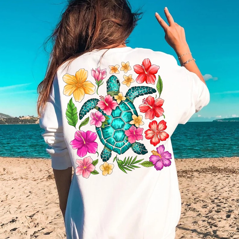 May include: White sweatshirt featuring a colourful graphic of a sea turtle surrounded by tropical flowers. The turtle is teal blue with a green shell and the flowers are pink, yellow, red, and purple.