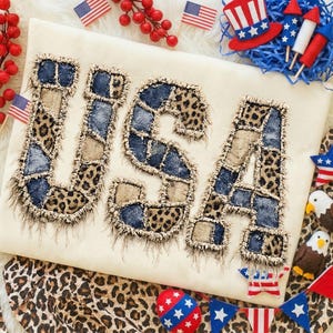 May include: A cream-coloured shirt featuring the word "USA" in patchwork lettering. The letters combine denim, leopard print, and tan fabrics. The image is decorated with miniature American flags and patriotic decorations.