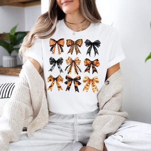 May include: White t-shirt featuring a grid of nine decorative bows in various styles and colors. The bows include leopard print, black lace, and Halloween-themed designs with bats and pumpkins. The shirt is a casual, everyday wear item.