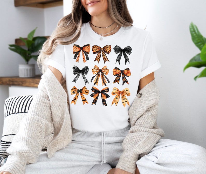 May include: White t-shirt featuring a grid of nine decorative bows in various styles and colors. The bows include leopard print, black lace, and Halloween-themed designs with bats and pumpkins. The shirt is a casual, everyday wear item.
