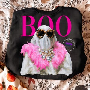May include: A black sweatshirt featuring a large pink graphic of a ghost wearing leopard print sunglasses and a pink feather boa with a crystal statement necklace. The word BOO is printed in large pink letters above the ghost figure.