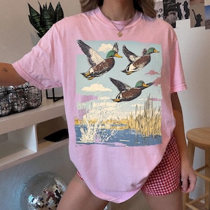 May include: A light pink t-shirt featuring a graphic of three ducks in flight over a body of water with reeds. The ducks are brown, white, and green, set against a blue sky with clouds. The water is blue with white splashes.