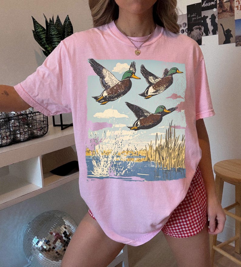 May include: A light pink t-shirt featuring a graphic of three ducks in flight over a body of water with reeds. The ducks are brown, white, and green, set against a blue sky with clouds. The water is blue with white splashes.