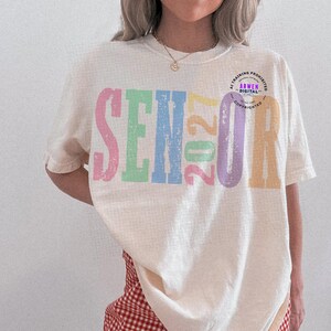 Puede incluir: Una camiseta de manga corta de color crema con el texto SENIOR 2027 en letras grandes, desgastadas y multicolores. Las letras son de color rosa, verde claro, azul, amarillo y morado. Un peque&ntilde;o logotipo circular es visible cerca del texto.