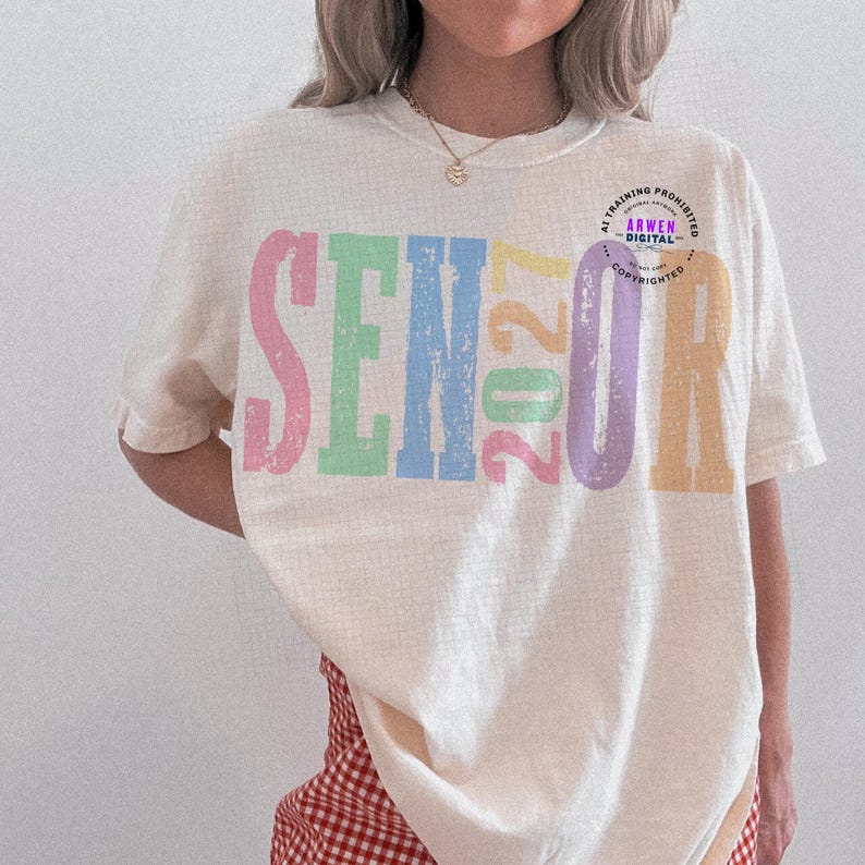 Puede incluir: Una camiseta de manga corta de color crema con el texto SENIOR 2027 en letras grandes, desgastadas y multicolores. Las letras son de color rosa, verde claro, azul, amarillo y morado. Un peque&ntilde;o logotipo circular es visible cerca del texto.