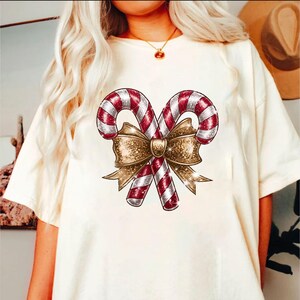 Puede incluir: Camiseta color crema con un dise&ntilde;o festivo de dos bastones de caramelo con rayas rojas y blancas, atados con un lazo dorado brillante. El gr&aacute;fico est&aacute; centrado en la camiseta, creando un tema navide&ntilde;o.