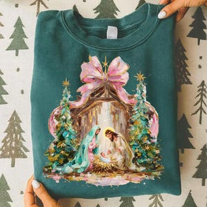 May include: Teal sweatshirt featuring a Nativity scene with a pink bow, Christmas trees, and figures of Mary, Joseph, and baby Jesus. The design is a colorful illustration with gold accents.