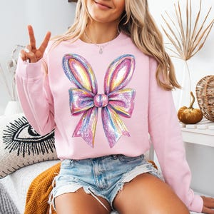 May include: A pink sweatshirt with a large iridescent bow graphic on the front. The bow is made up of multiple colors, including pink, purple, blue, and green.