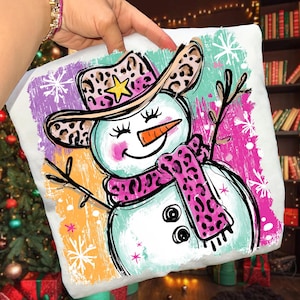 May include: A white square cushion with a playful snowman illustration. The snowman is adorned with a leopard print cowboy hat and scarf, a carrot nose, and twig arms. The background features colourful brushstrokes and snowflakes, evoking a festive winter atmosphere.