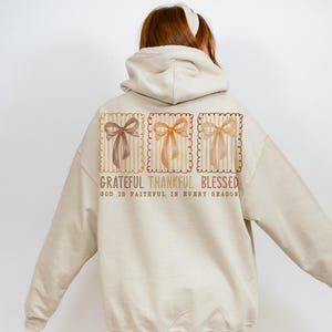 May include: Beige hoodie with a graphic on the back featuring three bow illustrations in brown, orange, and yellow. The text reads "Grateful Thankful Blessed" with "God is faithful in every season" below.