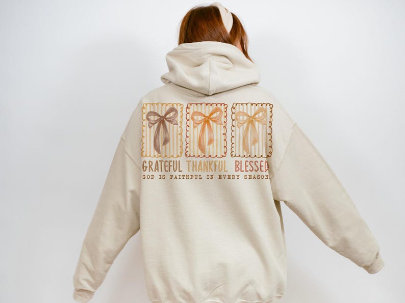 May include: Beige hoodie with a graphic on the back featuring three bow illustrations in brown, orange, and yellow. The text reads "Grateful Thankful Blessed" with "God is faithful in every season" below.