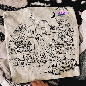 May include: A square beige pillow cover featuring a black line art illustration of a ghost, a church, corn stalks, and pumpkins. A sign in the foreground reads LIVE HERE NOW. The fabric has a subtle grid pattern and a circular watermark logo.