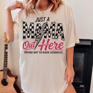 Puede incluir: Camiseta blanca con un dise&ntilde;o a cuadros blanco y negro y un rayo rosa. El texto "Just a Mama Out Here Trying Not to Raise Assholes" est&aacute; impreso en la camiseta.