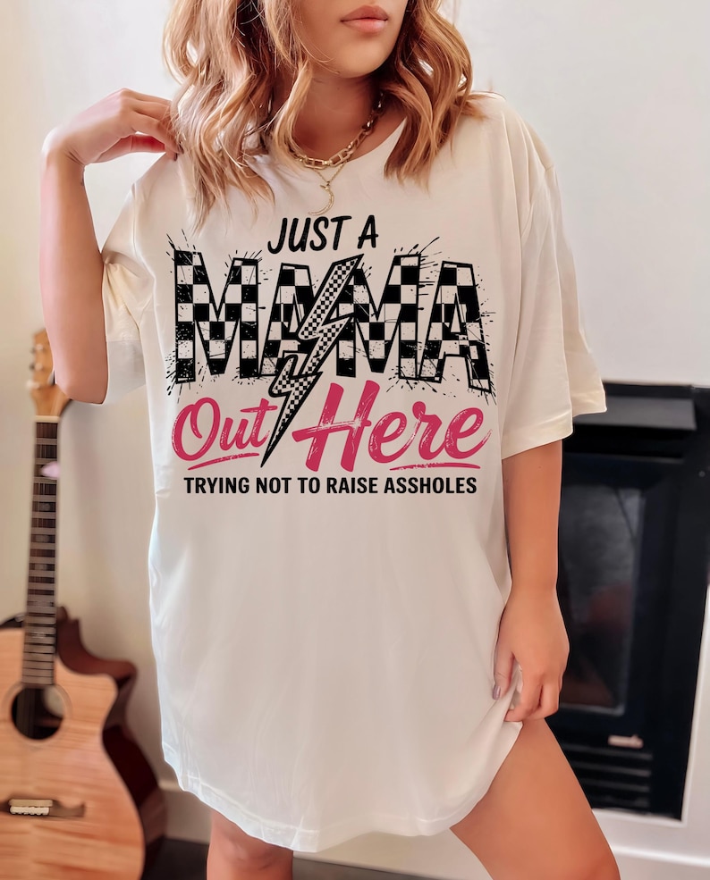 Puede incluir: Camiseta blanca con un dise&ntilde;o a cuadros blanco y negro y un rayo rosa. El texto "Just a Mama Out Here Trying Not to Raise Assholes" est&aacute; impreso en la camiseta.