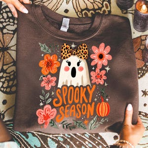 K&ouml;nnte beinhalten: Ein braunes Sweatshirt mit einem Geisterdesign, das eine Schleife mit Leopardenmuster tr&auml;gt, umgeben von orangefarbenen und rosa Blumen und den Worten "Spooky Season" in Orange. Ein K&uuml;rbis ist ebenfalls im Design enthalten. Das Sweatshirt ist gefaltet.