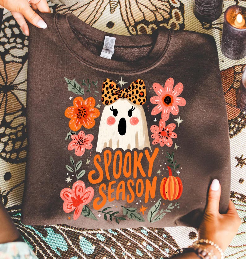 K&ouml;nnte beinhalten: Ein braunes Sweatshirt mit einem Geisterdesign, das eine Schleife mit Leopardenmuster tr&auml;gt, umgeben von orangefarbenen und rosa Blumen und den Worten "Spooky Season" in Orange. Ein K&uuml;rbis ist ebenfalls im Design enthalten. Das Sweatshirt ist gefaltet.