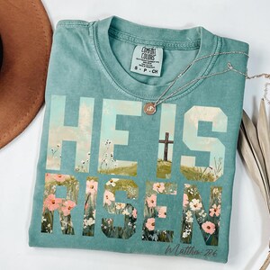 May include: A teal blue t-shirt with the words "He is Risen" in large white letters. The letters are filled with a painting of a field of flowers and a cross. The text "In Matthew 28:6" is printed in white below the letters.