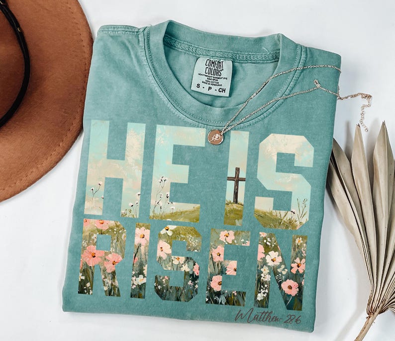 May include: A teal blue t-shirt with the words "He is Risen" in large white letters. The letters are filled with a painting of a field of flowers and a cross. The text "In Matthew 28:6" is printed in white below the letters.