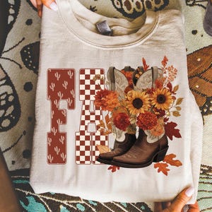 May include: A cream-colored sweatshirt featuring the word "FA" in a western-style font with cactus and checkered patterns. The design includes brown cowboy boots, sunflowers, and fall foliage. The overall theme is autumn and country.