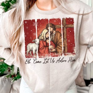 May include: Beige sweatshirt featuring a religious illustration with a family, lamb, and cross on a red background. The text "Oh Come Let Us Adore Him" is printed below the image. The sweatshirt is a neutral color.