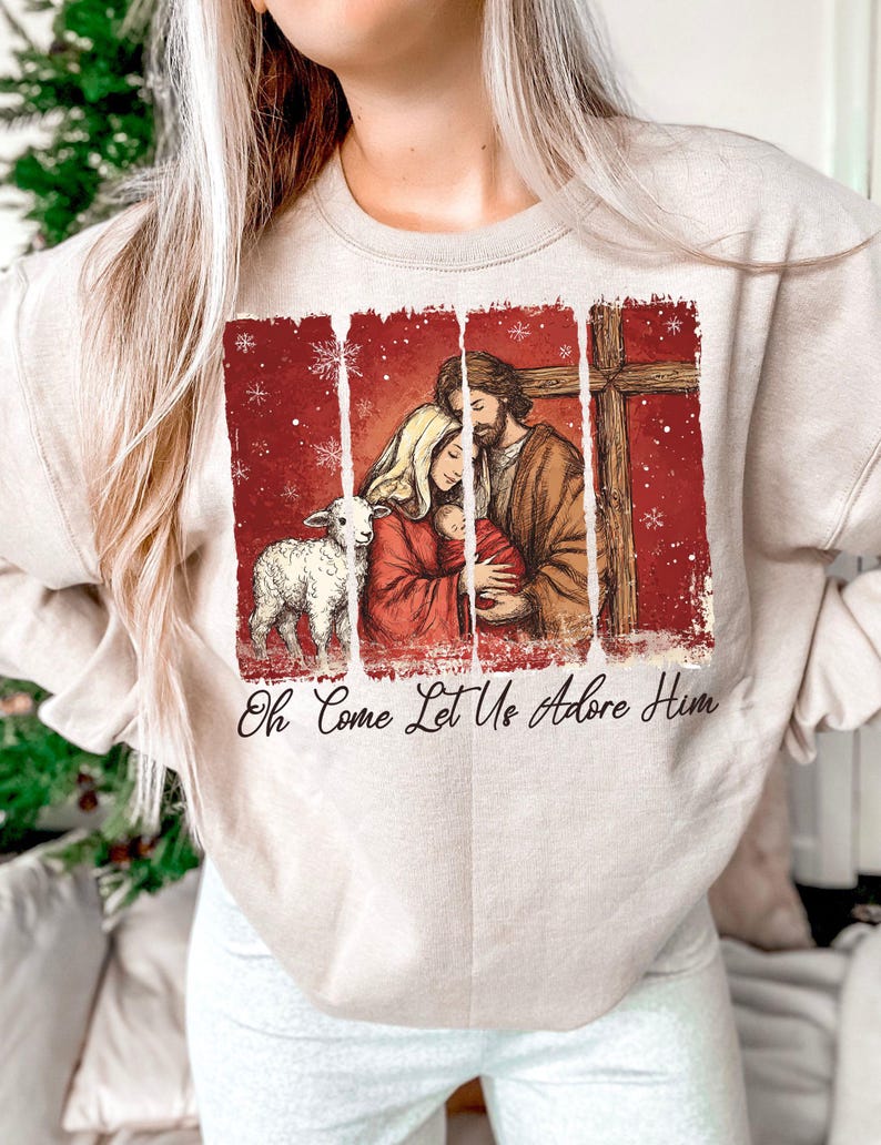 May include: Beige sweatshirt featuring a religious illustration with a family, lamb, and cross on a red background. The text "Oh Come Let Us Adore Him" is printed below the image. The sweatshirt is a neutral color.