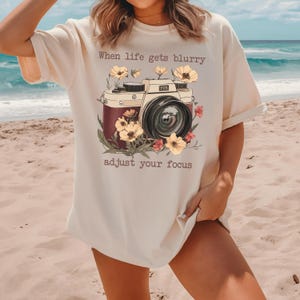May include: A white t-shirt with a vintage camera graphic and the text "When life gets blurry adjust your focus".