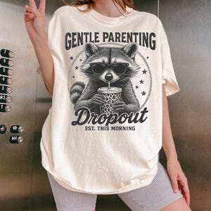 Puede incluir: Camiseta color crema con un gr&aacute;fico de mapache con gafas de sol y sosteniendo una bebida. El texto "GENTLE PARENTING Dropout EST. THIS MORNING" est&aacute; impreso encima y debajo de la ilustraci&oacute;n del mapache. La camiseta es de corte holgado.