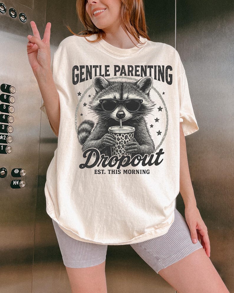 Puede incluir: Camiseta color crema con un gr&aacute;fico de mapache con gafas de sol y sosteniendo una bebida. El texto "GENTLE PARENTING Dropout EST. THIS MORNING" est&aacute; impreso encima y debajo de la ilustraci&oacute;n del mapache. La camiseta es de corte holgado.